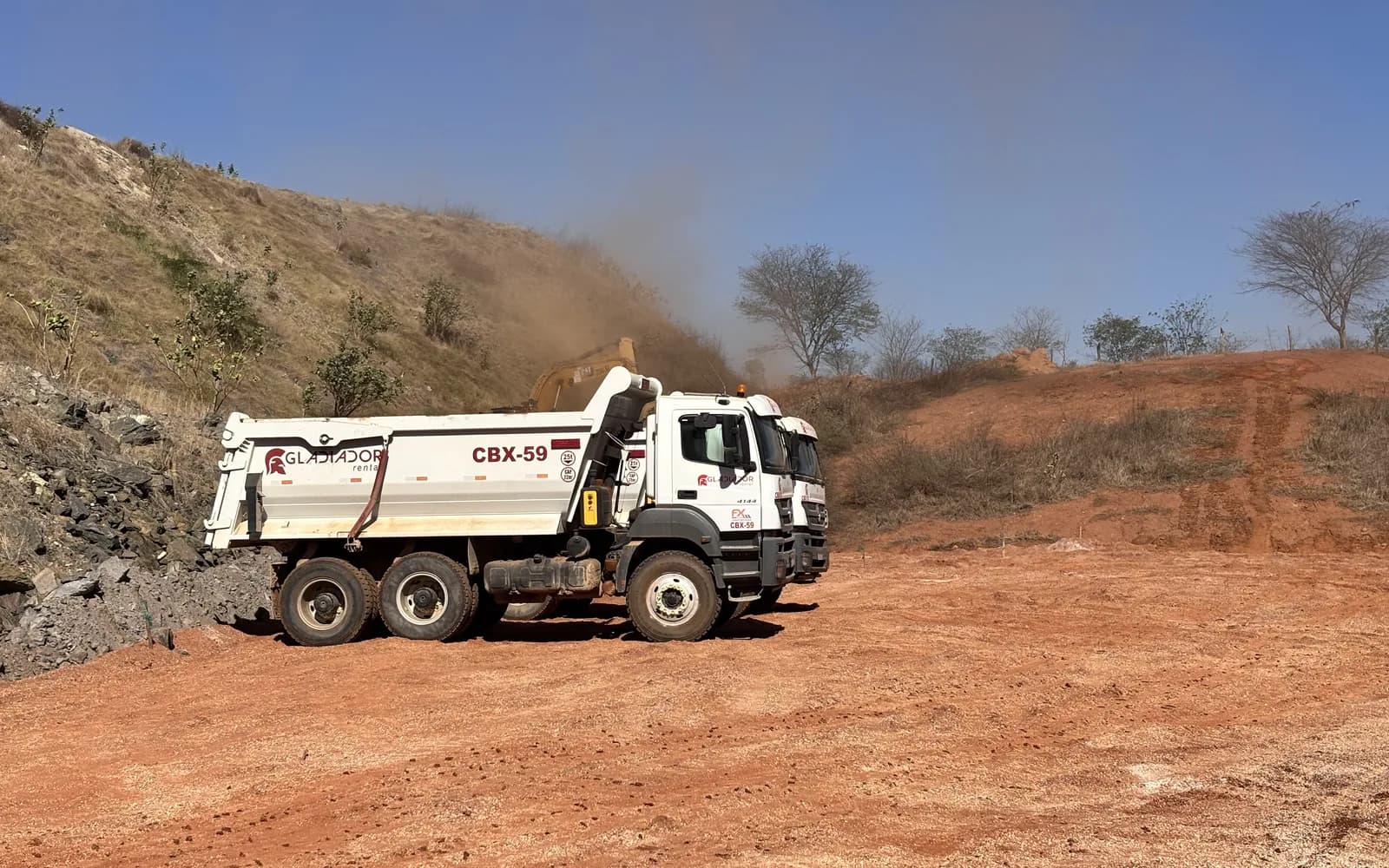 Caminhão basculante da Gladiador, empresa do Grupo FX Minas, parado em praça de terra vermelha seca enquanto a poeira sobe da escavadeira ao fundo, a cena que o controle de poeira na mineração precisa resolver