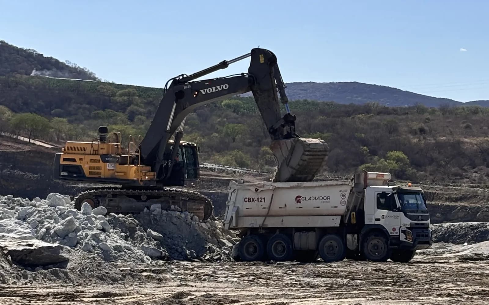 Escavadeira em cima da pilha de rocha despejando a caçamba na báscula de um basculante da Gladiador, empresa do Grupo FX Minas, parado no piso de terra da praça de carga